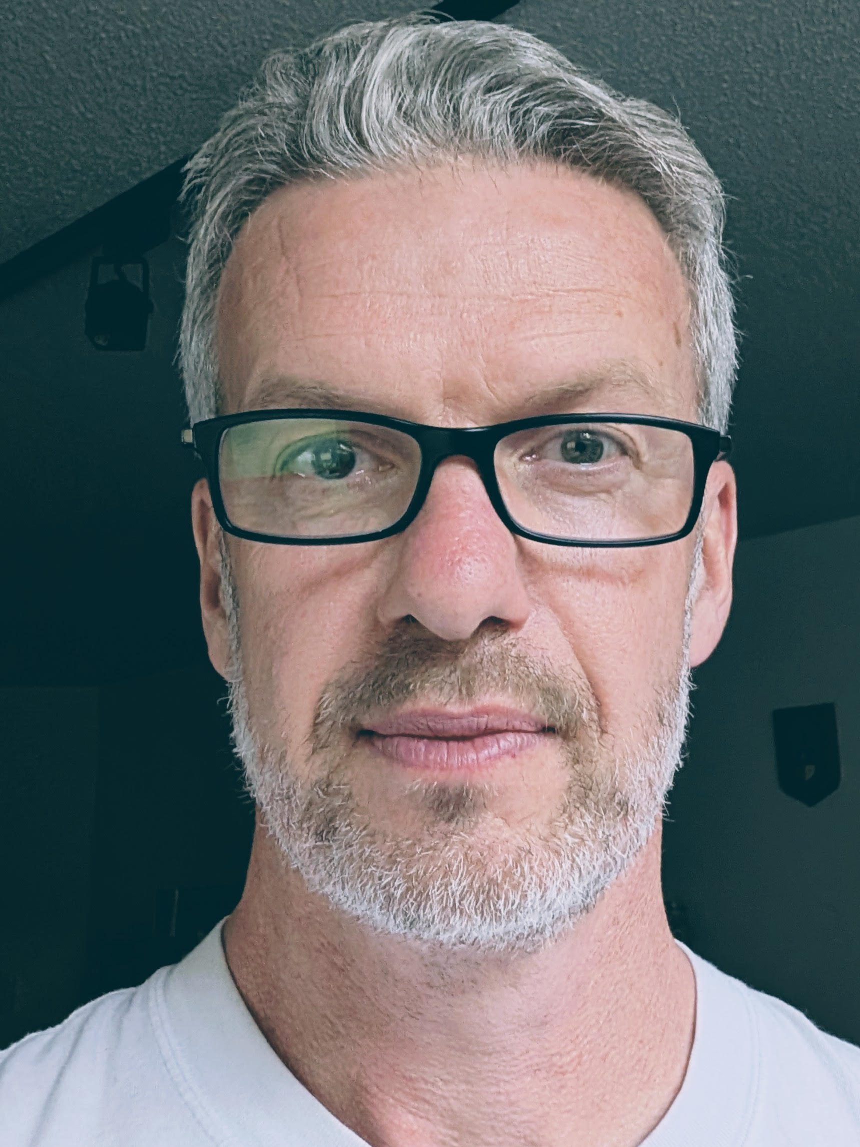 Close-up portrait of a man with gray hair and a beard, wearing glasses and a white shirt, in a softly lit room.
